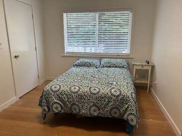 Das kleine, minimalistische Schlafzimmer hat ein Doppelbett mit einer bunt gemusterten Bettdecke, einen weißen Nachttisch, ein großes Fenster mit Jalousien und einen Holzboden.