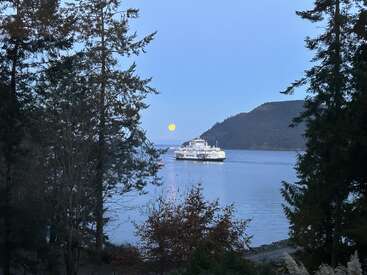 Eine Fähre fährt über eine ruhige blaue Bucht, umrahmt von hohen immergrünen Bäumen. Der Vollmond geht über den fernen Hügeln auf und sorgt für eine friedliche, malerische Abendstimmung.