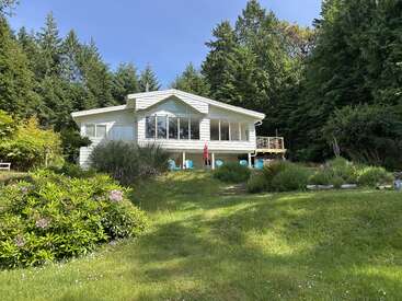 Inmitten von üppigem Grün und Bäumen steht ein charmantes weißes Haus mit großen Fenstern, einer geräumigen Terrasse, blauen Stühlen und einem friedlichen, sonnenbeschienenen Garten, der es umgibt.