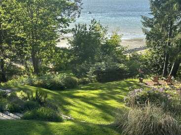 Eine ruhige Aussicht auf das Meer mit üppigem grünen Rasen, Blumenbeeten, hohen Bäumen und Adirondack-Stühlen mit Blick auf einen ruhigen Strand und blaues Wasser an einem sonnigen Tag.