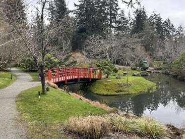 Eine ruhige Gartenszene mit einer leuchtend roten Brücke über einen friedlichen Teich, umgeben von Bäumen, gewundenen Wegen, grünem Gras und einem immergrünen Wald im Hintergrund.