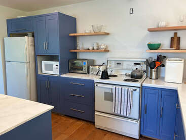 Diese gemütliche Küche hat blaue Schränke, weiße Geräte, Holzregale, ein gestreiftes Handtuch auf dem Ofen und verschiedene Utensilien, Geschirr und Kleingeräte, die fein säuberlich angeordnet sind.