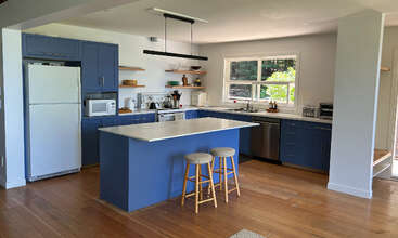 Diese moderne Küche hat blaue Schränke, eine weiße Arbeitsinsel mit zwei Hockern, Edelstahlgeräte, offene Holzregale, ein großes Fenster und einen Holzfußboden. Ein heller, einladender Raum.