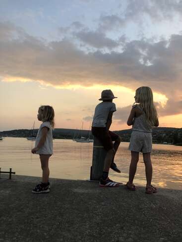 Trois enfants se tiennent près d'un front de mer paisible au coucher du soleil. Des voiliers flottent à l'arrière-plan. Le ciel resplendit de couleurs chaudes, créant une scène calme et pittoresque.
