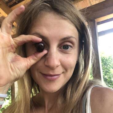 A smiling woman with long hair holds a dark cherry in front of her left eye, making a playful gesture. Natural light and greenery are visible outside.