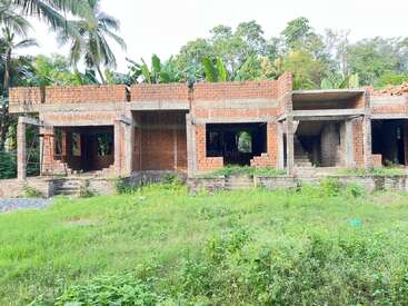An unfinished brick building stands amidst lush greenery. Concrete columns and beams support the structure, surrounded by palm trees and banana plants in the background.