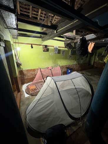 A dimly lit rustic room with an open wooden roof, green walls, hanging bags, two mosquito nets covering mats, and a single bulb illuminating the area.