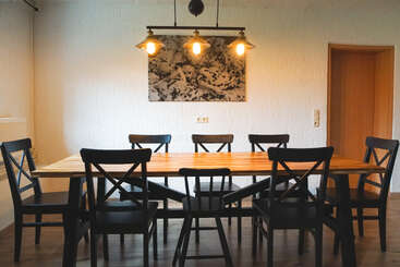 Un comedor moderno con una larga mesa de madera rodeada de ocho sillas negras, una cálida iluminación colgante, paredes blancas con textura y una decoración minimalista con una única obra de arte abstracta.
