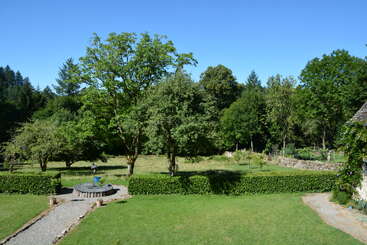 A sunny garden scene with green lawns, neat shrubbery, gravel paths, and tall leafy trees, surrounded by a stone wall and dense forest background.