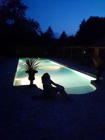 A person sits beside a glowing pool at dusk, surrounded by silhouettes of trees. The tranquil scene exudes peacefulness, highlighted by gentle pool lighting and shadows.
