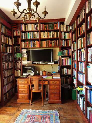 A cozy home library with wooden shelves filled with books surrounds a wooden desk and chair. A chandelier hangs above. A TV and green lamps present.