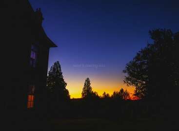 Un hermoso cielo al atardecer pasa del amarillo al azul intenso. Las siluetas de los árboles y una casa con ventanas iluminadas crean una atmósfera serena y tranquila al atardecer.
