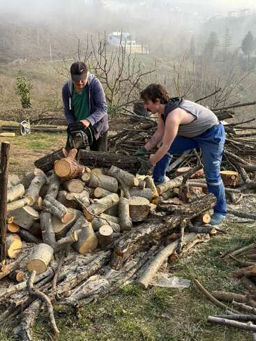 Zwei Typen schneiden und stapeln draußen Brennholz. Einer benutzt 'ne Kettensäge, während der andere die Holzscheite ordnet. Im Hintergrund sieht man Nebel, kahle Bäume und Häuser in der Ferne.