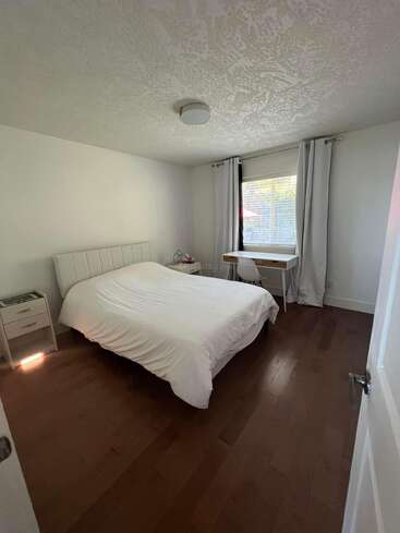 Esta habitación tiene una cama con ropa de cama blanca, mesilla de noche y un escritorio junto a la ventana con cortinas grises y suelos de madera, creando un aspecto limpio y minimalista.