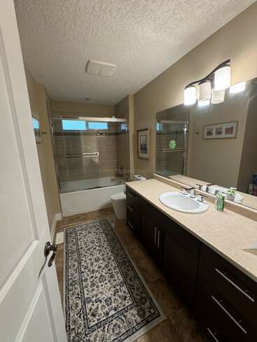 Esta imagen muestra un cuarto de baño moderno con lavabo doble, espejo grande, bañera con puertas de cristal, alfombra estampada, paredes de color beige, suelo de baldosas y obras de arte en la pared.