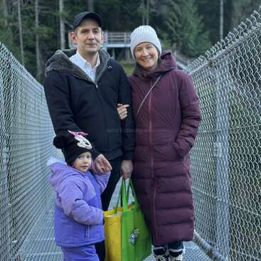 Una familia de tres miembros, bien abrigados, permanecen juntos en un puente rodeado de vallas metálicas y árboles. Sonríen, cogidos de la mano y llevando una bolsa verde.