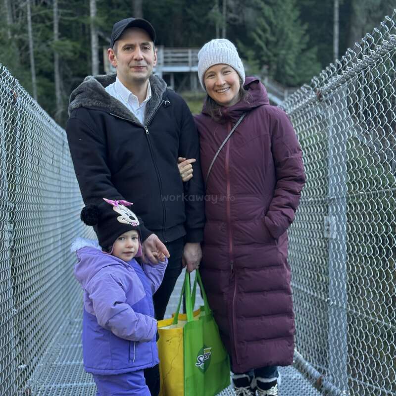 Una familia de tres miembros, bien abrigados, permanecen juntos en un puente rodeado de vallas metálicas y árboles. Sonríen, cogidos de la mano y llevando una bolsa verde.