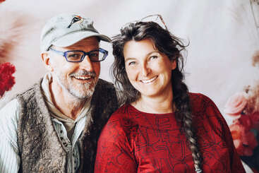Un homme souriant avec des lunettes et une casquette se tient à côté d'une femme joyeuse aux longs cheveux tressés et à la chemise rouge à motifs. Tous deux semblent heureux et détendus.
