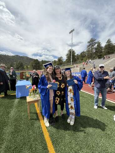 Deux diplômés, vêtus de toges et de casquettes bleues, se tiennent aux côtés d'une femme vêtue d'une robe en forme de tournesol. Ils sont souriants et tiennent leurs diplômes, sur un terrain de sport pendant la remise des diplômes.