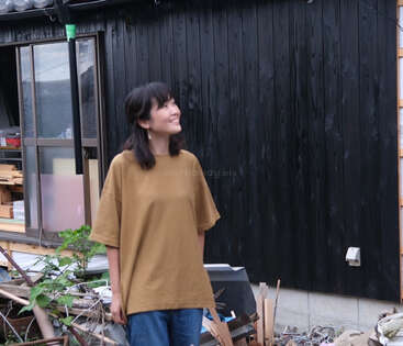 A woman in a loose brown shirt and blue jeans stands smiling outside a black wooden house, surrounded by scattered debris and green plants, looking upward contentedly.