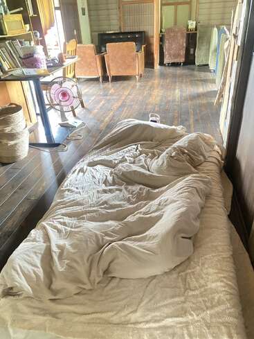 A cozy futon bed with crumpled blankets lies on a wooden floor in a sunlit, rustic room with bookshelves, chairs, and a standing fan nearby.