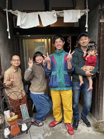 Five people, including a child, stand happily in front of a doorway. Four adults smile and flash peace signs, while the child is held gently.