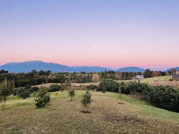 Un paisaje sereno al atardecer, con campos verdes, pequeños árboles dispersos, bosques lejanos, cadenas montañosas en el horizonte, casas de madera y un cielo azul rosado pastel.