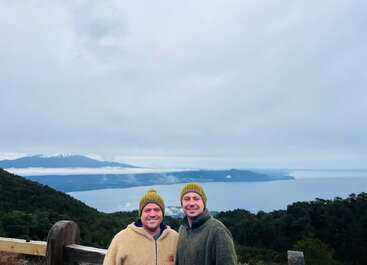Dos personas vestidas con ropa de abrigo y sombreros verdes a juego sonríen juntas, de pie frente a un paisaje de montañas, bosques y un gran lago tranquilo.