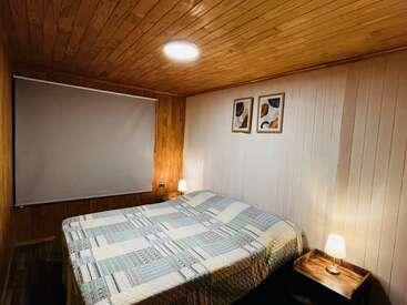 Este acogedor dormitorio cuenta con una cama grande con una colcha de retazos, dos mesillas de noche con lámparas, paredes y techo con paneles de madera y dos obras de arte abstracto.