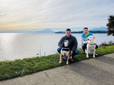 Dos hombres sonríen arrodillados junto a un lago con dos perros. Las montañas nevadas y el cielo nublado crean un paisaje de fondo. La hierba y la acera enmarcan el apacible momento.