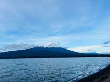 Un lago sereno se extiende por el primer plano, reflejando el cielo. Una montaña nevada se alza majestuosa en el centro, bajo un vasto cielo azul con nubes difusas.