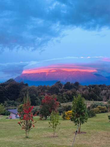 Una vibrante puesta de sol proyecta un resplandor rosado sobre una montaña coronada de nubes. Abajo, exuberantes campos verdes y árboles de colores crean un paisaje tranquilo y pintoresco.