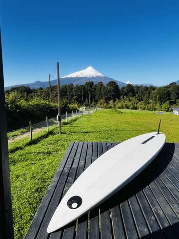 Una tabla de surf blanca descansa sobre una cubierta de madera con vistas a un campo de un verde vibrante, con un volcán nevado y un cielo azul despejado al fondo.