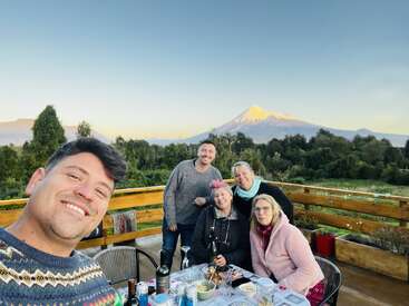 Cinco personas disfrutan de una comida al aire libre en una terraza de madera, sonrientes con bebidas y comida. Detrás de ellos, un volcán nevado se eleva sobre un frondoso bosque verde.
