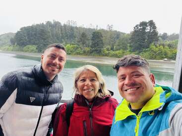Tres personas sonríen para una foto al aire libre cerca de un lago, con chaquetas de colores. Detrás de ellos hay árboles, colinas verdes y un cielo nublado, que crean una atmósfera serena.