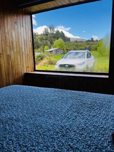 Una acogedora habitación de madera cuenta con una cama revestida de azul frente a una gran ventana, con una vista panorámica de un coche blanco, verdes colinas, árboles y un cielo azul.