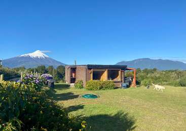 Eine gemütliche Holzhütte steht auf einem üppigen grünen Rasen, umgeben von Blumen, mit einem schneebedeckten Vulkan und Bergen im Hintergrund unter einem klaren Himmel.