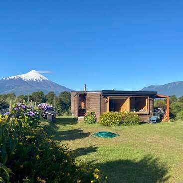 A cozy wooden cabin sits on a lush green lawn, surrounded by flowers, with a snow-capped volcano and mountains in the background under a clear sky.