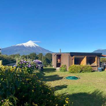 Une cabane en bois confortable est posée sur une pelouse verdoyante, entourée de fleurs, avec un volcan aux sommets enneigés et des montagnes en arrière-plan, sous un ciel dégagé.