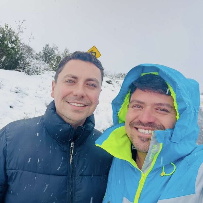 Zwei Männer lächeln in einer verschneiten Umgebung im Freien, beide tragen Winterjacken. Es schneit, hinter ihnen sind Bäume und ein Straßenschild zu sehen, die eine fröhliche Winterszene schaffen.