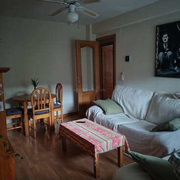 A cozy living room with a white couch, green pillows, colorful tablecloth, wooden dining set, open door, ceiling fan, and monochrome wall art.