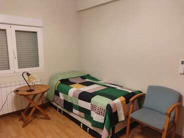 This room has a single bed with a patchwork quilt, a light blue armchair, wooden floors, a round table, a lamp, and a window.