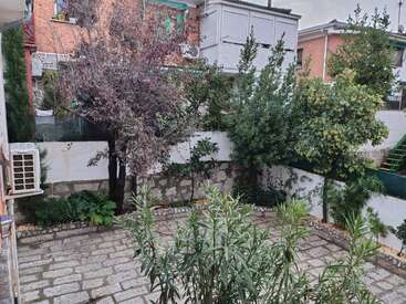A small, paved courtyard features various trees and shrubs along the stone wall. Surrounding buildings overlook the garden. An air conditioning unit is mounted on the left.