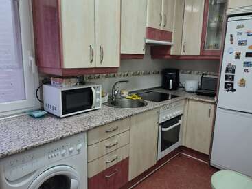 This is a compact kitchen with beige and red cabinets, a granite countertop, washing machine, microwave, fridge with magnets, oven, sink, toaster, and kettle.
