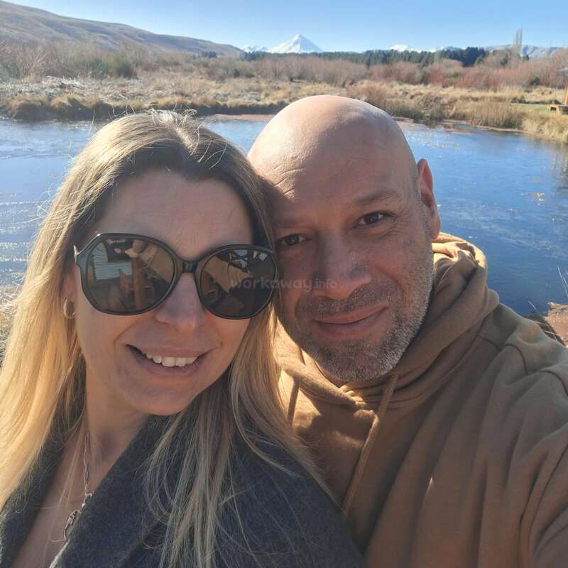 Ein lächelndes Paar posiert für ein Selfie im Freien an einem ruhigen Teich, mit Bergen und einem schneebedeckten Gipfel im sonnigen Hintergrund. Beide scheinen glücklich zu sein.