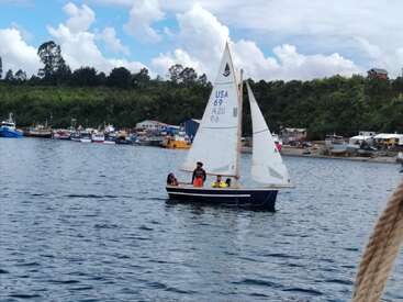 Four people sail a small boat marked "USA 69" on calm blue water, with a green shoreline, moored boats, and houses visible in the background.