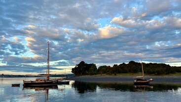 Two sailboats float peacefully on calm water near a shore lined with trees. The evening sky is covered with scattered clouds, reflecting beautifully on the water.