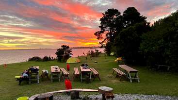 The image captures a vibrant sunset over a lakeside campsite, featuring picnic tables, scattered belongings, several yellow tents, lush trees, and a dramatic, colorful sky.