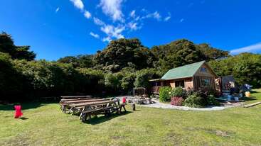 A cozy wooden cabin with a green roof sits surrounded by lush greenery. Wooden picnic tables, benches, and bright blue skies create a peaceful, inviting outdoor setting.