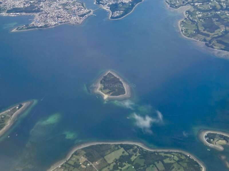 This aerial photo shows a small, heart-shaped island surrounded by blue water, with nearby landmasses featuring visible patches of green fields and developed areas.